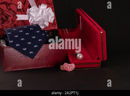 Anniversary gift concept. Silver pearl necklace in red box, candy wih blue tie and decorated gift box on black background Stock Photo