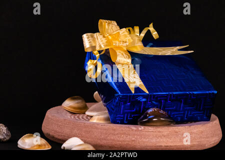 Blue gift box decorated with golden ribbon on Stone and spreaded seashells on dark background. Gift concept Stock Photo