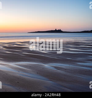 Beautiful patterns in the sand at Embleton Bay reflect the pre-sunrise sky with the imposing ruins of Dunstanburgh Castle on the horizon. Stock Photo