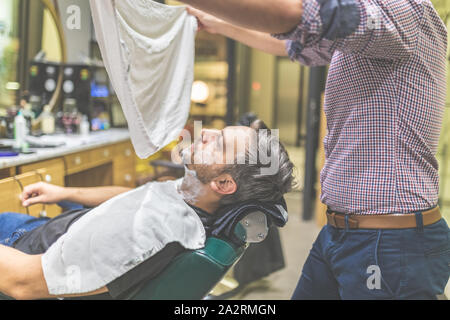 Barber covering client with towel before giving him a shave at barbershop. Stock Photo