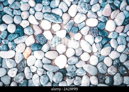 A full-frame background texture featuring a dense arrangement of smooth, rounded blue, grey, and white pebbles on dry sand. Stock Photo