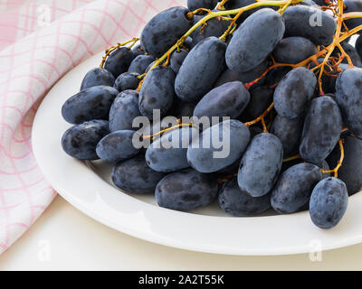 Sweet and tasty blue grapes with long berries on a white plate on a table. Bunches of juicy ripe dark grapes. Close-up of delicious dessert fruits. Stock Photo