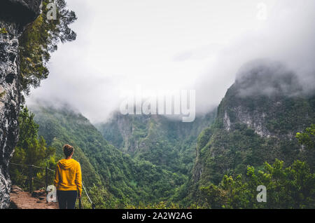 Young woman in yellow sweatshirt on a viewpoint in Levada Caldeirao Verde, Madeira, Portugal. Green scenic mountains in fog, misty landscape. Female traveler. Instagram, hipster filter. Stock Photo