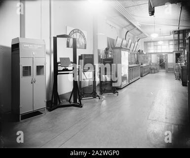 Glass Negative, Showroom, circa 1930s, Black and white full plate glass negative of the interior of a Kodak Australasia Pty Ltd retail showroom - a wide wooden room, walls lined with counters, and display cabinets. In the foreground are several large pieces of Kodak equipment for use by professional photographers - a Kodak Roll Film Dryer and large studio plate cameras. A woman is visible inside the lift shaft at the rear, right of Stock Photo