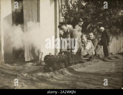 Digital Photograph - Man & Two Boys Riding Miniature Train for Charity, Sandringham, 1943-1944, A man and two boys riding in a miniature train in a backyard. Two men are standing behind and a shed is visible to the left. This photograph shows a miniature train ride for the 'Bomb Children of Britain' charity, Sandringham, circa 1943. The three men riding the train are unidentified but the man behind on the left is Robert Edwards, who built the train. The man on the right is his father Stock Photo