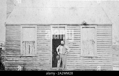 Negative - Man Standing in Cottage Doorway, Western Australia (?), circa 1890, A bearded man, holding a hat, standing in the doorway of a small Victorian timber cottage. There are two large windows on either side of him, from which curtains can be seen. The roof of the cottage is made of iron, and a brick chimney is located on the left Stock Photo