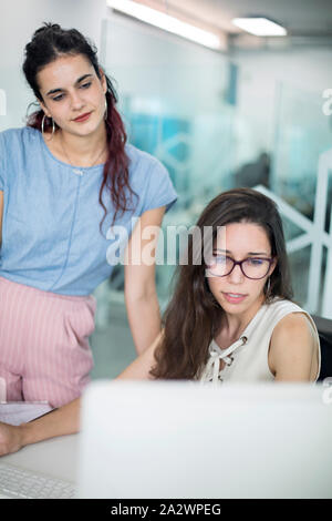 Two young women watching messages on computer screen, in a coworking space. Modern office interior with glass divisions for freelance workers. Stock Photo