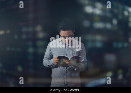 Male student wearing long sleeves and eye glasses reading a book peacefully inside an isolated dark room at night time. Wide view shot. Stock Photo