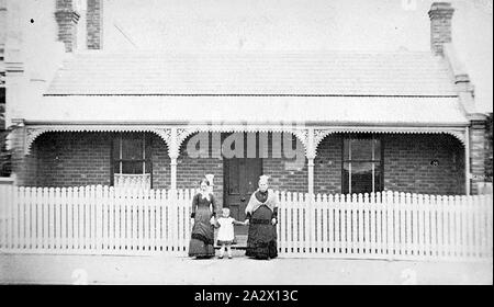 Negative - North Melbourne, Victoria, circa 1885, Two women and a small child stand in front of their brick house. This house has a verandah decorated with iron lacework and brick chimneys. A picket fence runs along the front of their property Stock Photo