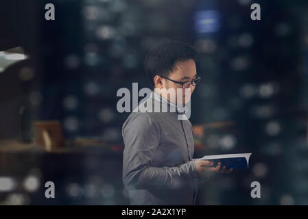 Side view close up shot of a young male student wearing long sleeves and glasses reading a book peacefully and isolated inside his office room. Stock Photo