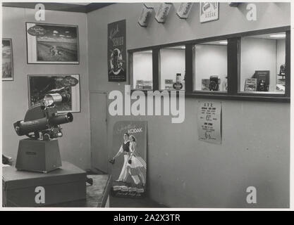 Photograph, Exhibition Stand, Chamber of Manufactures Exhibition, Adelaide, Mar - May 1947, Black and white photograph of a section of the Kodak Australasia Pty Ltd exhibition stand or trade show booth at the Chamber of Manufactures Exhibition held from March to May 1947 in Adelaide. This view focuses on the wall of recessed display cabinets with products and films arranged within. There is a 'Go with a Kodak' poster on the ground, as well as posters for the Australian Photo Review Stock Photo