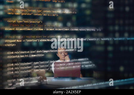 Blurred male computer programmer on the background, thinking while programming or coding using a laptop inside an office at night with reflected codes Stock Photo