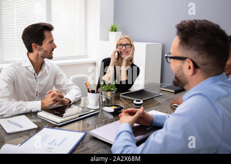 Young Business Woman Speaking On The Phone Sitting With Colleagues In A Meeting Stock Photo