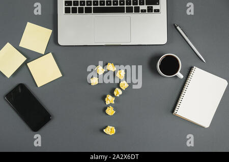 Yellow question mark lined with crumpled paper balls, notebook, cup coffee, phone, pen on gray background. Asking, Decisions. FAQ concept. Concept ide Stock Photo