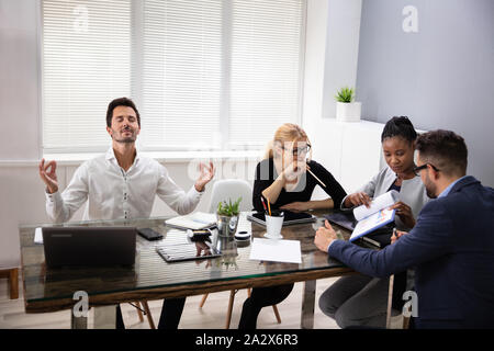 Young Male Office Worker Meditating At Workplace Ignoring Stressful Meeting With Colleagues Stock Photo
