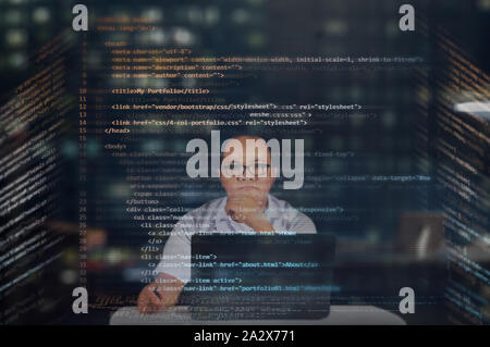 Young IT male student reading his HTML codes projected on a virtual hologram screen with his laptop on the desk. Thinking and focused working at night Stock Photo