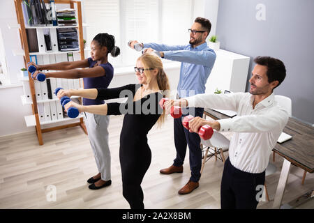 Happy Young Businesspeople Standing In Row Exercising With Dumbbells In Office Stock Photo