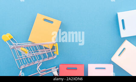 Abstract design element, annual sale, shopping season concept, mini yellow cart with colorful paper bag on pastel blue background, top view, flat lay Stock Photo