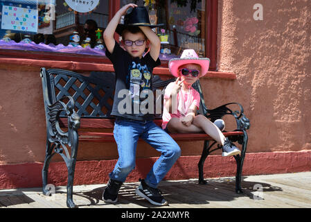 A young boy adjust his black cowboy hat and little girl in pink cowboy hat and pink sunglasses flashes a peace sign, sitting on a bench before a store Stock Photo