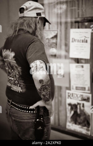 Rear view of long straggly haired middle aged man wearing baseball cap and knife on belt and arm tattoos studies storefront covered in posters Stock Photo