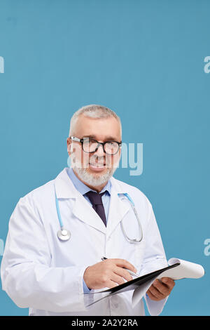 Portrait of mature doctor with grey hair and in eyeglasses smiling at camera while standing and writing a prescription over blue background Stock Photo