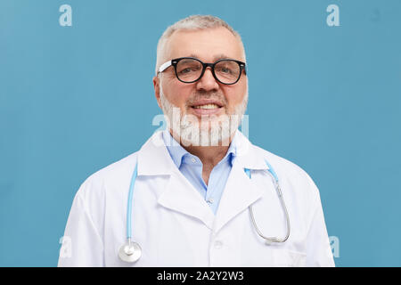 Portrait of mature doctor with grey hair and beard wearing eyeglasses and white coat smiling at camera isolated on blue background Stock Photo