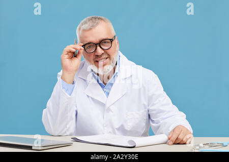 Portrait of mature doctor with grey hair and beard and in eyeglasses smiling at camera while working with documents at the table Stock Photo