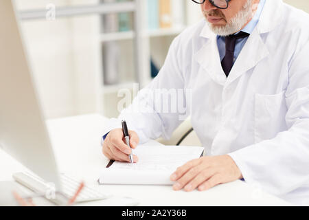 Close-up of senior doctor in white coat sitting at the table and writing in notepad during his work day at office Stock Photo