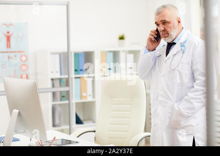 Serious senior doctor in white coat talking on mobile phone while standing at office Stock Photo