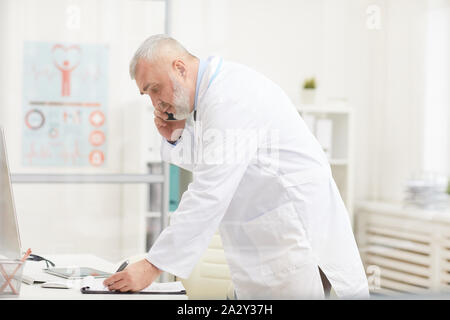 Senior doctor in white coat concentrating on his work he talking on mobile phone and making notes in document at office Stock Photo