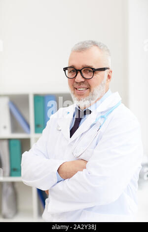 Portrait of senior doctor in eyeglasses and in white coat standing with his arms crossed and smiling at camera at office Stock Photo