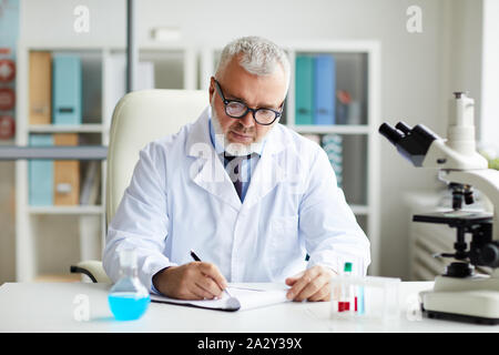 Senior scientist in eyeglasses sitting at the table writing in document while working with test tubes and microscope at the laboratory Stock Photo