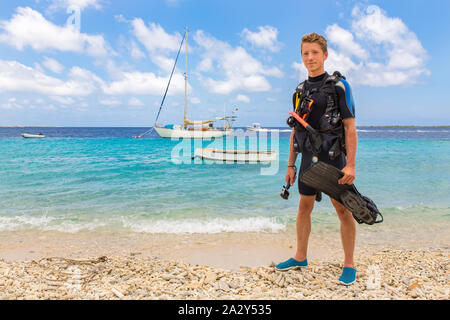 Caucasian male diver stands on beach of Bonaire with sea and boats Stock Photo