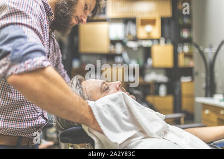 Barber covering client with towel before giving him a shave at barbershop. Stock Photo