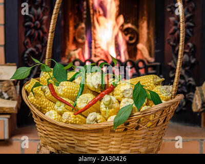 A wicker basket to the top filled with ears of ripe corn and hot peppers. In the background firewood burning in the fireplace insert. Cozy rustic inte Stock Photo