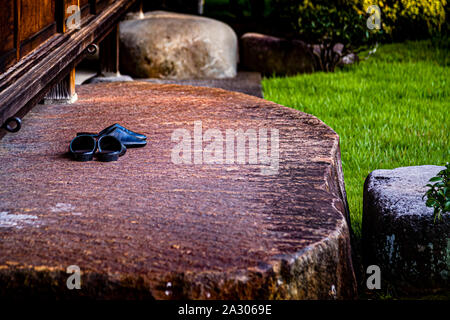 Black lacquered slippers sit neatly side by side on a massive stone slab at the entrance of Ryokan Sanyoso in Izunokuni, Japan, in the morning shadow of the surrounding trees. The estate was built in 1929 for the family of Mitsubishi founder Yataro Iwasaki, became a ryokan in 1947, and has been an important cultural property since 2017. Stock Photo