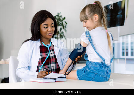 young and cute female African doctor taking a blood pressure to a little girl. Pediatrician measuring blood pressure of little girl patient Stock Photo
