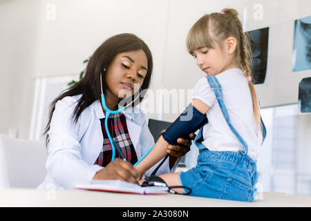 Little Caucasian girl at paediatrician office measuring blood pressure. Young African woman doctor measuring blood pressure
