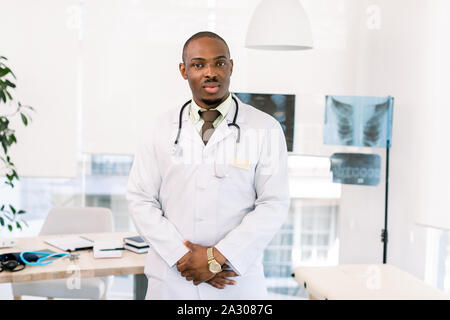 Young handsome friendly african american man doctor in white coat standing in modern medical office. Light modern clinic, radiography on the Stock Photo