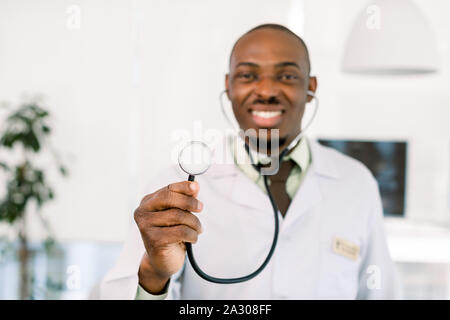 Young handsome smiling African medical physician doctor man holding stethoscope, over modern hospital background. Focus on stethoscope Stock Photo