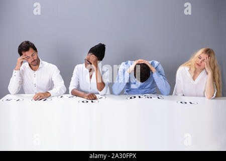 Tired Stressful Multiracial Judges Sitting On Chair With Scoring Point On Table Against Wall Stock Photo