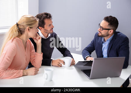 Happy Young Couple Consulting Male Friendly Financial Advisor With Coffee Cups At Office Desk Stock Photo