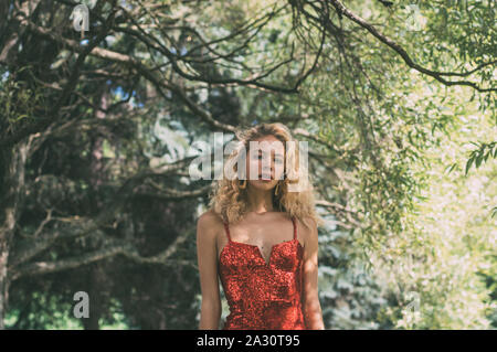 Portrait of beautiful blond girl in red short dress with sequins in park Stock Photo