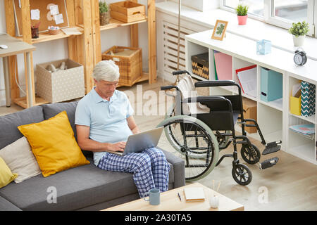 Serious senior man using laptop while resting on sofa with wheelchair near by him in the living room Stock Photo