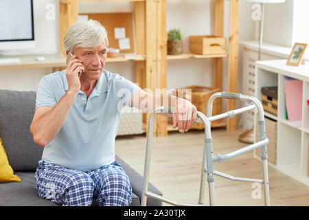Senior man with grey hair sitting on sofa leaning on the walker and talking on mobile phone in the room Stock Photo