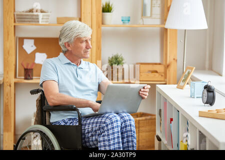 Senior disabled man in wheelchair looking out the window while using laptop computer at home Stock Photo