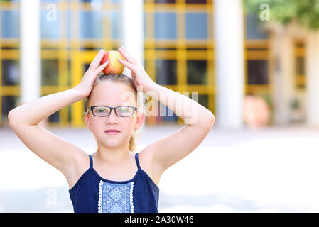 Happy Girl In Eyeglasses With Apple on Her Head. Great Portrait Of School Pupil Outside Classroom. Back to school, balance, success, target and healthy eating for child concept. Stock Photo