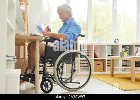 Senior disabled man sitting in wheelchair and examining business documents at the table in front of computer in domestic room Stock Photo