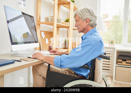 Senior disabled businessman sitting in wheelchair at the table and looking at computer monitor with financial graphics while working at office Stock Photo