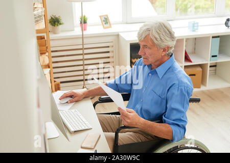 Serious disabled senior man sitting at his workplace in front of computer monitor and working with documents at home Stock Photo